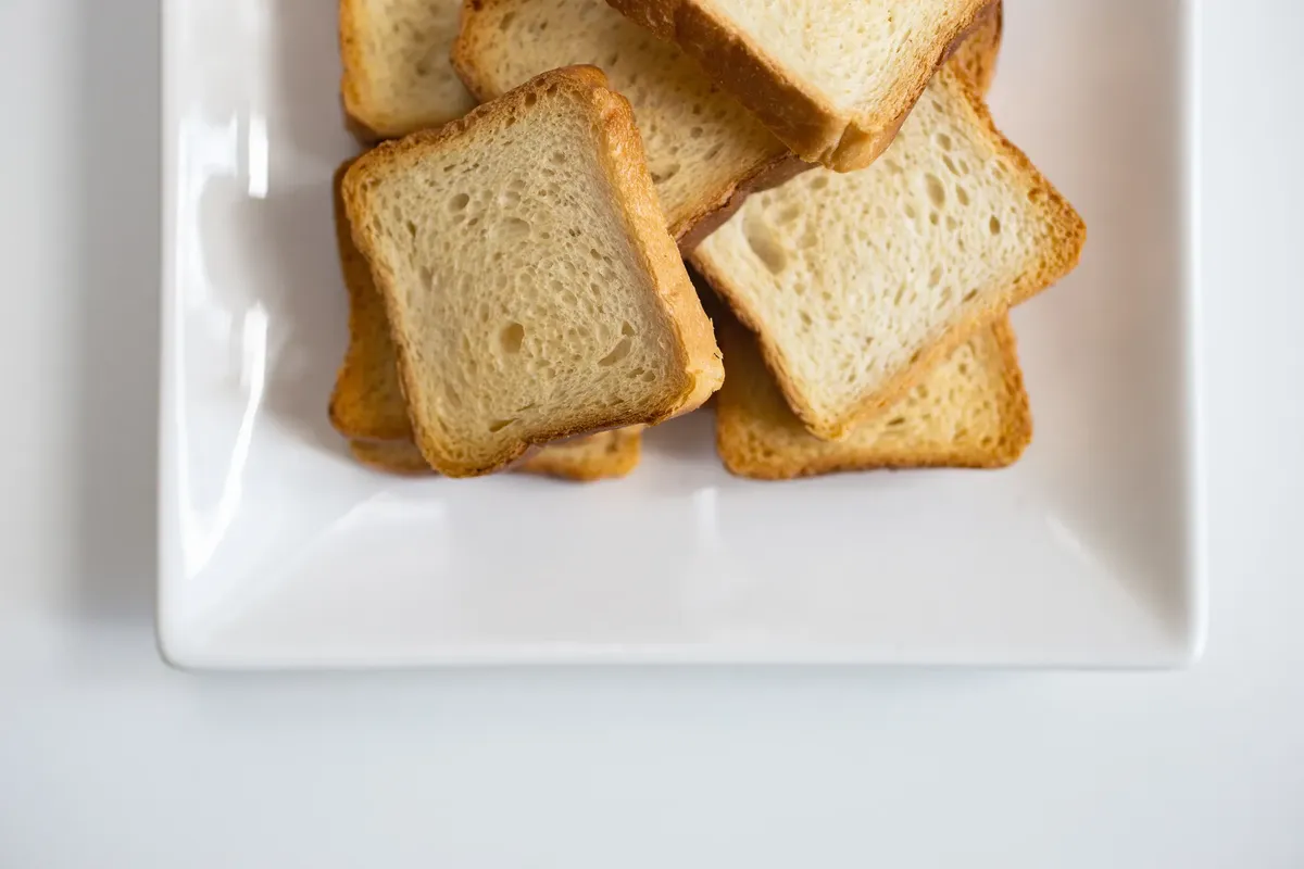 Stack of toasted bread slices on a white ceramic plate, perfect for breakfast or snacks.